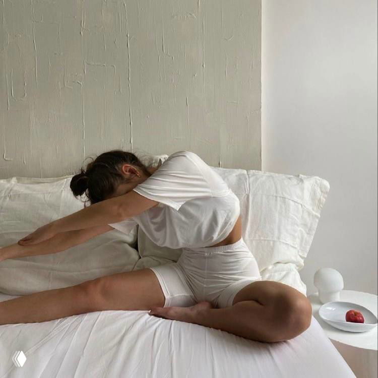 Человек делает растяжку на кровати в светлой комнате: спортивный комплект, мягкая постель и минималистичный интерьер создают спокойную атмосферу утренней практики.