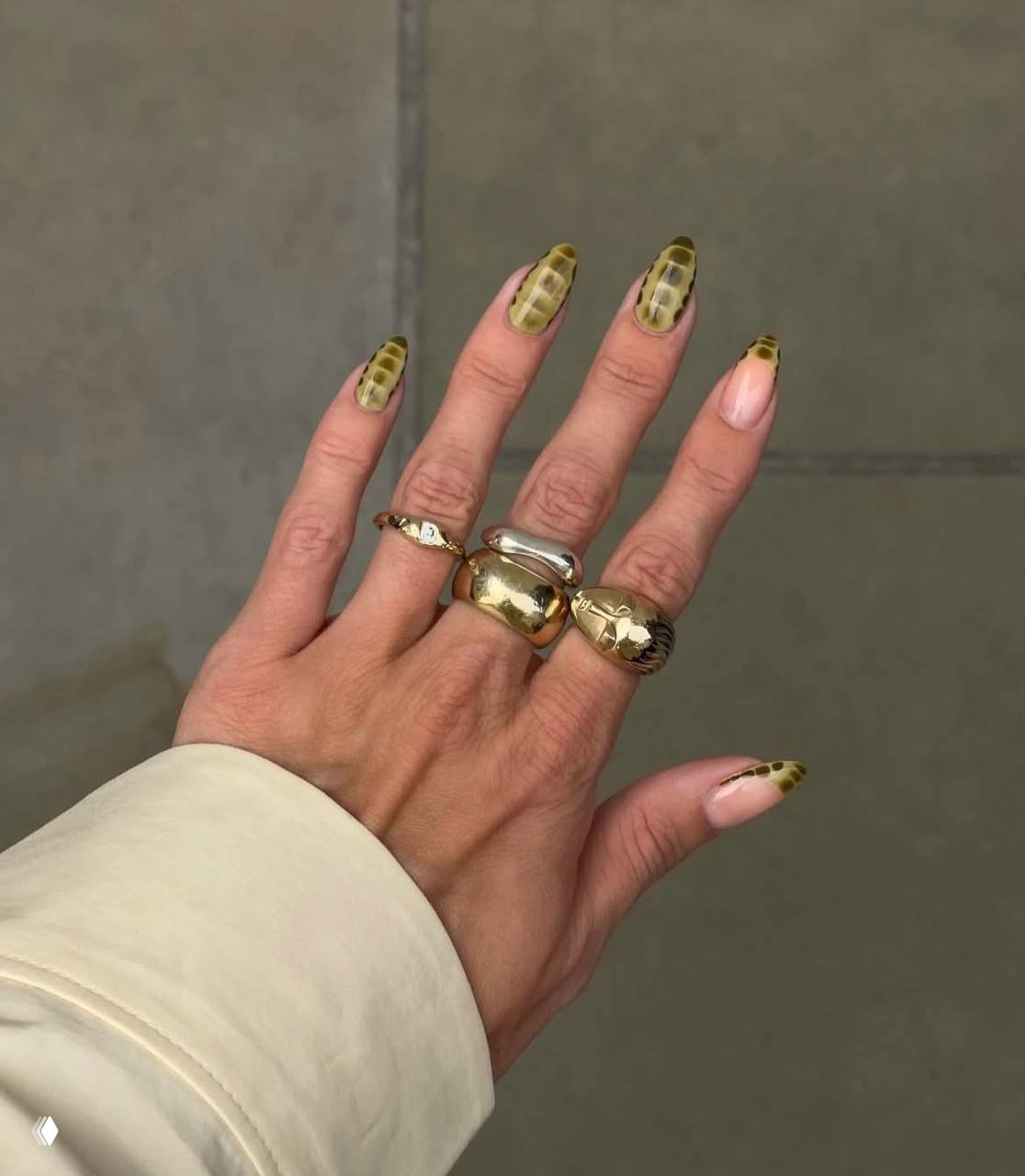 Рука с кольцами демонстрирует анималистичный маникюр под змеиную кожу — зелёно-оливковые чешуйчатые мотивы на длинных ногтях, нейтральный фон.