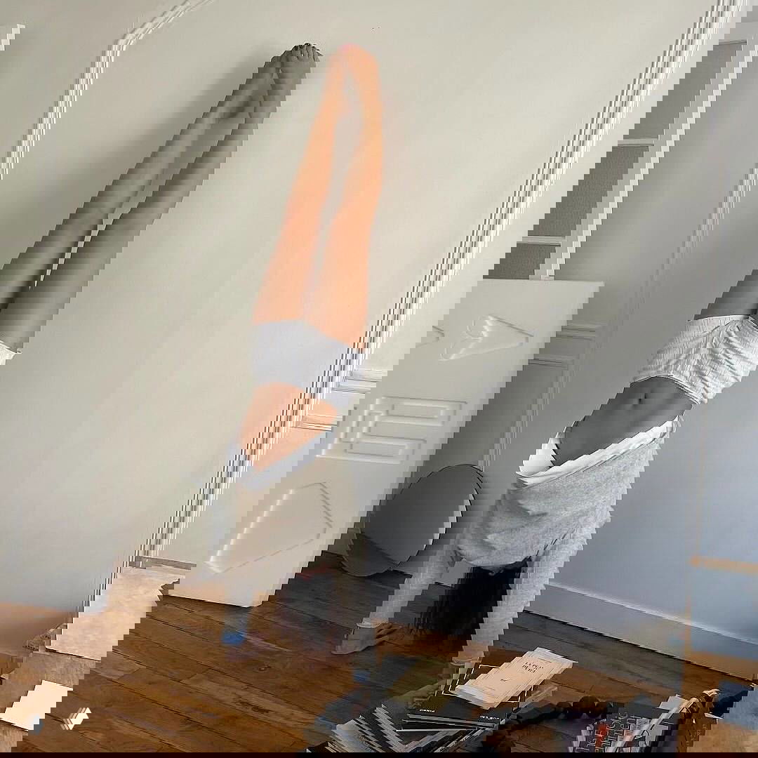 Человек стоит в стойке на руках у стены в комнате с деревянным полом и белыми стенами; кадр передаёт баланс, лёгкую акробатику и домашнюю атмосферу.