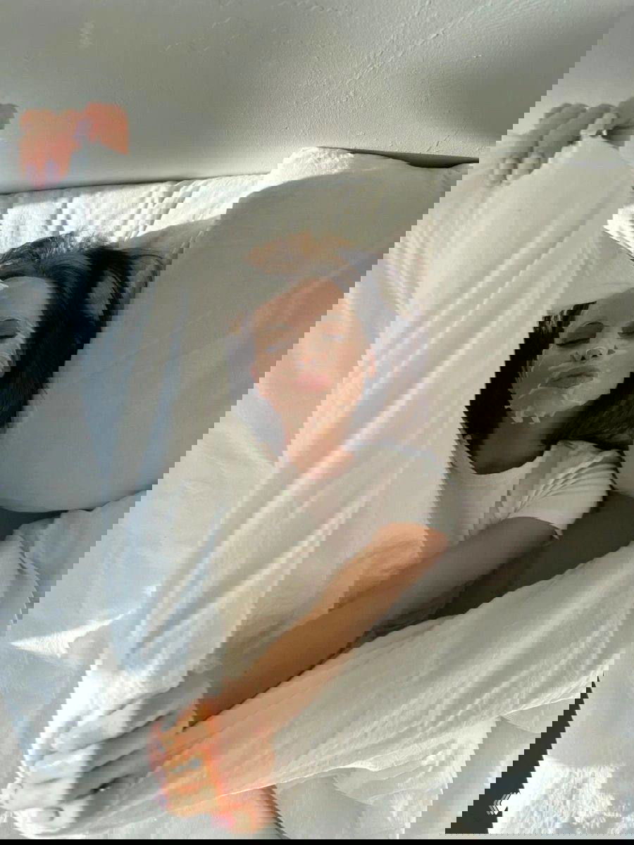 Девушка лежит в постели в тканевой маске и тянется из-под одеяла; сцена показывает использование маски утром после ночи в домашней спальне.