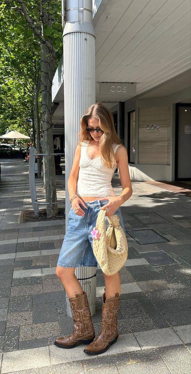 Уличный кадр: девушка в белом топе, джинсовых шортах и коричневых казаках стоит на тротуаре — сочетание ковбойских сапог и летнего лука.
