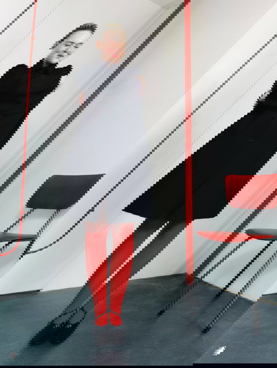 Фотосессия: женщина в черном пальто и ярких красных колготках стоит в студии рядом с красным стулом, полный рост, стилизованный интерьер.