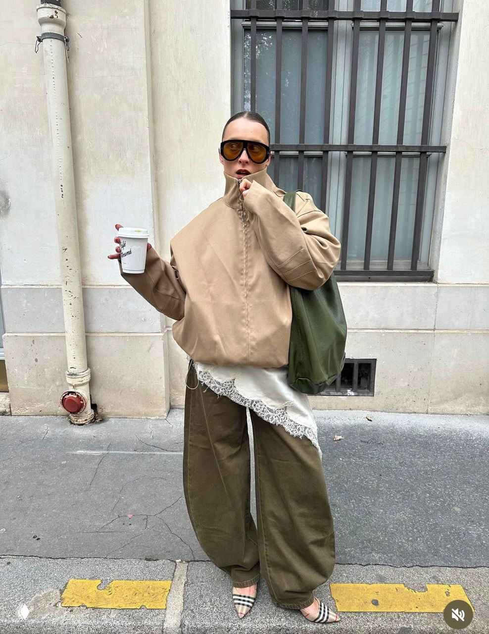 Женский лук в бежевой куртке с высоким воротником-стойкой, кружевная нижняя деталь и широкие зелёные брюки; модель держит стакан кофе на улице.