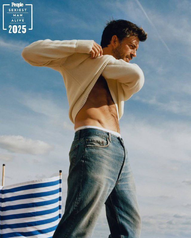 Джонатан Бейли снимает свитер на открытом воздухе в кадре фотосессии People 2025: видно плечо, торс, джинсы и небесный фон.