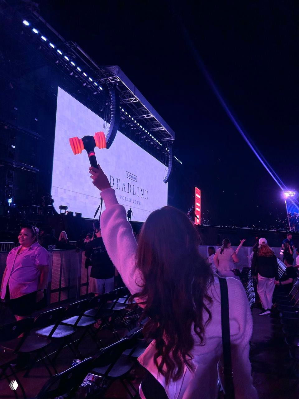 Зрительница держит светящийся лайтстик на фоне большой сцены и рекламного экрана; видны ряды кресел и вечерняя подсветка.