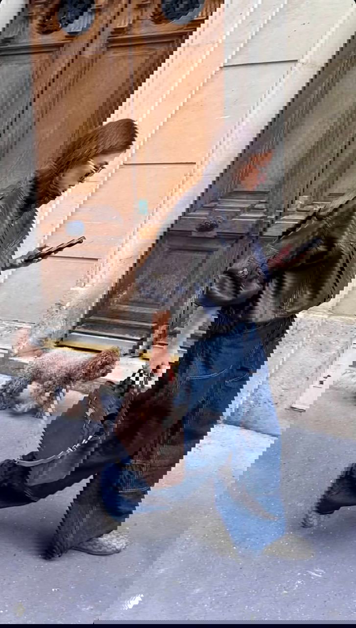 Девушка в тёмной кожаной куртке с высоким воротником-стойкой идёт по тротуару с сумкой и небольшим пуделем на поводке, динамичный городской кадр.