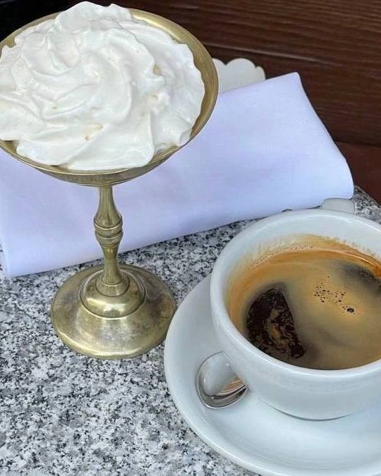 Кофейная чашка эспрессо на белой тарелке рядом с блюдом со взбитыми сливками, кадр со столом в кафе и мягким естественным освещением.