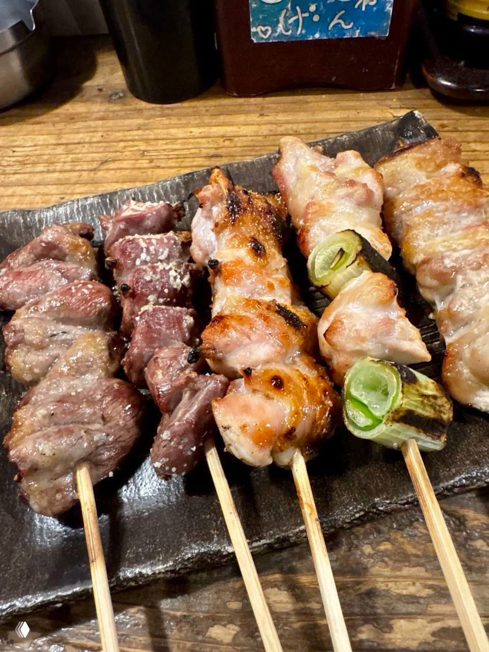 Тарелка с несколькими шпажками якитори разного вида: поджаренные кусочки мяса, лук и морепродукты на тёмной керамической посуде.