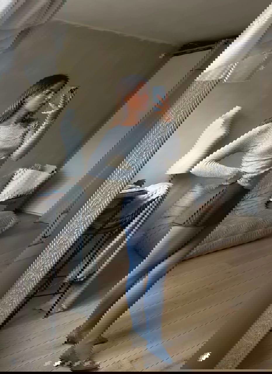 Селфи в зеркале: девушка примеряет образ в тёмно-серых плотных колготках и короткой юбке в комнате с манекеном и диваном, примерочная атмосфера.