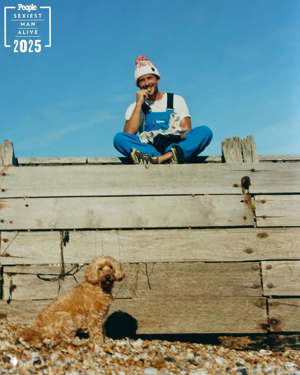 Джонатан Бейли сидит на деревянной набережной в шапке и комбинезоне, ниже на галечном пляже находится маленькая рыжая собака; композиция кадра широкая.