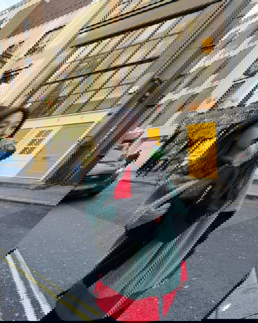 Девушка в зелёно-чёрной крупной пушистой куртке пьёт из бутылки на городской улице; сочетание объёмной верхней одежды и повседневной улицы.