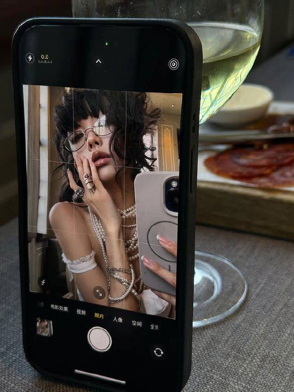 Селфи на экране телефона в чёрном чехле: девушка в очках и жемчужных украшениях, на заднем плане бокал и тарелка с закуской в кафе.