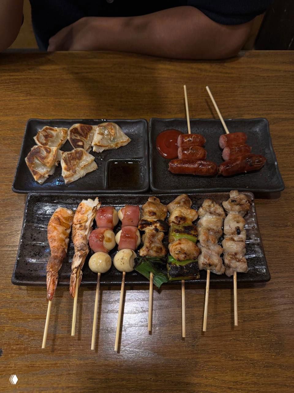 Несколько тарелок с закусками: гёдза, сосиски на шпажках и ряд якитори, поданные на деревянном столе в ресторане.