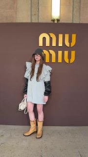 Модель в сером мини‑платье у стены с логотипом Miu Miu; образ дополнен чёрной панамой, жёлтыми сапогами и светлой сумкой в интерьере показа.