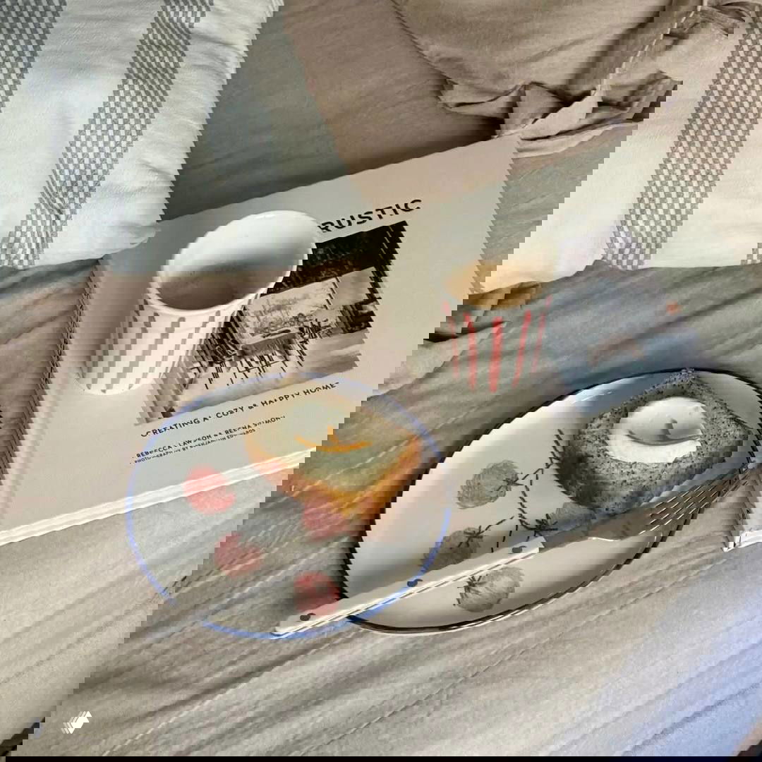 Завтрак на кровати: кусочек кекса с сыром, чашка кофе и книга на подложке — уютная сценка для ленивого утра и простых удовольствий.