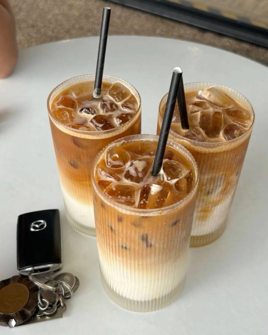Три высоких стакана айс-латте с яркими слоями молока и кофе и черными трубочками, стоящие на светлой поверхности рядом с ключами от машины.