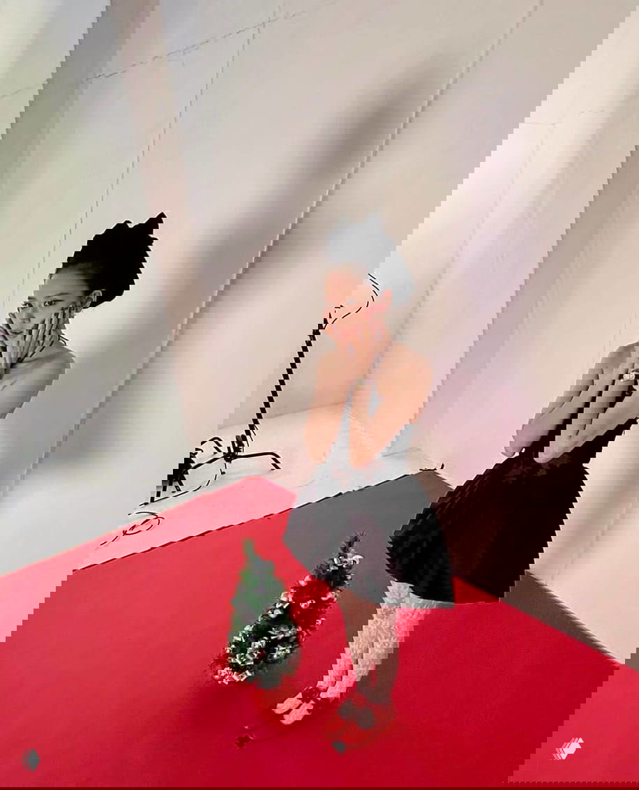 Айдол в чёрном мини-платье и банте стоит на красном фоне рядом с маленькой новогодней ёлочкой; ретро-эстетика образа и чулки в комплекте.