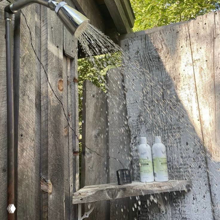 Уличный душ у деревянного забора: струя воды, флаконы с косметикой на полке и солнечные блики — простая деревенская летняя сцена.