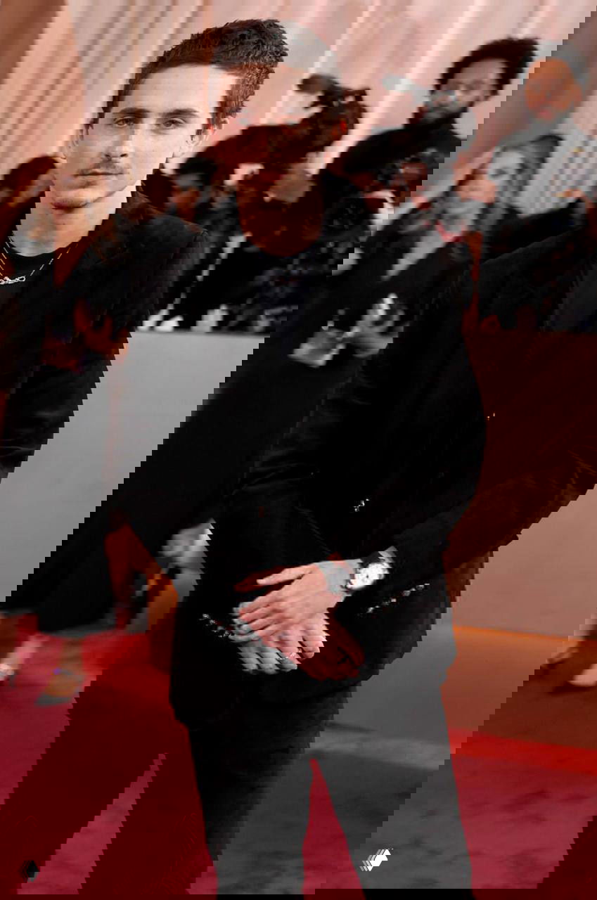 Мужчина в чёрном костюме стоит на красной ковровой дорожке; позади видны фотографы и гости, снимающие образ в полный рост.