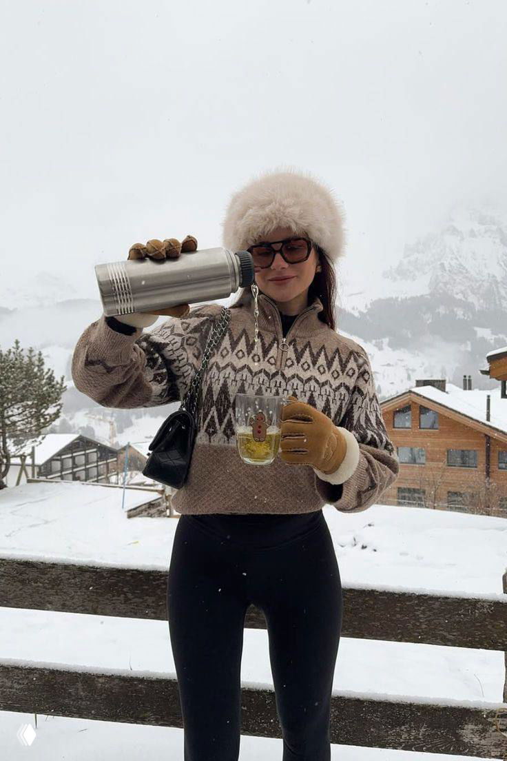 Девушка в узорчатом свитере и меховой шапке наливает горячий напиток на фоне заснеженных гор и деревянных шале, уютный зимний лук.