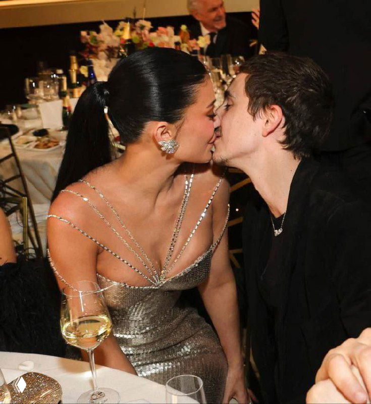 Двое людей целуются за столом на праздничном приёме; женщина в блестящем платье, на столе бокал и сервировка, атмосфера торжества.