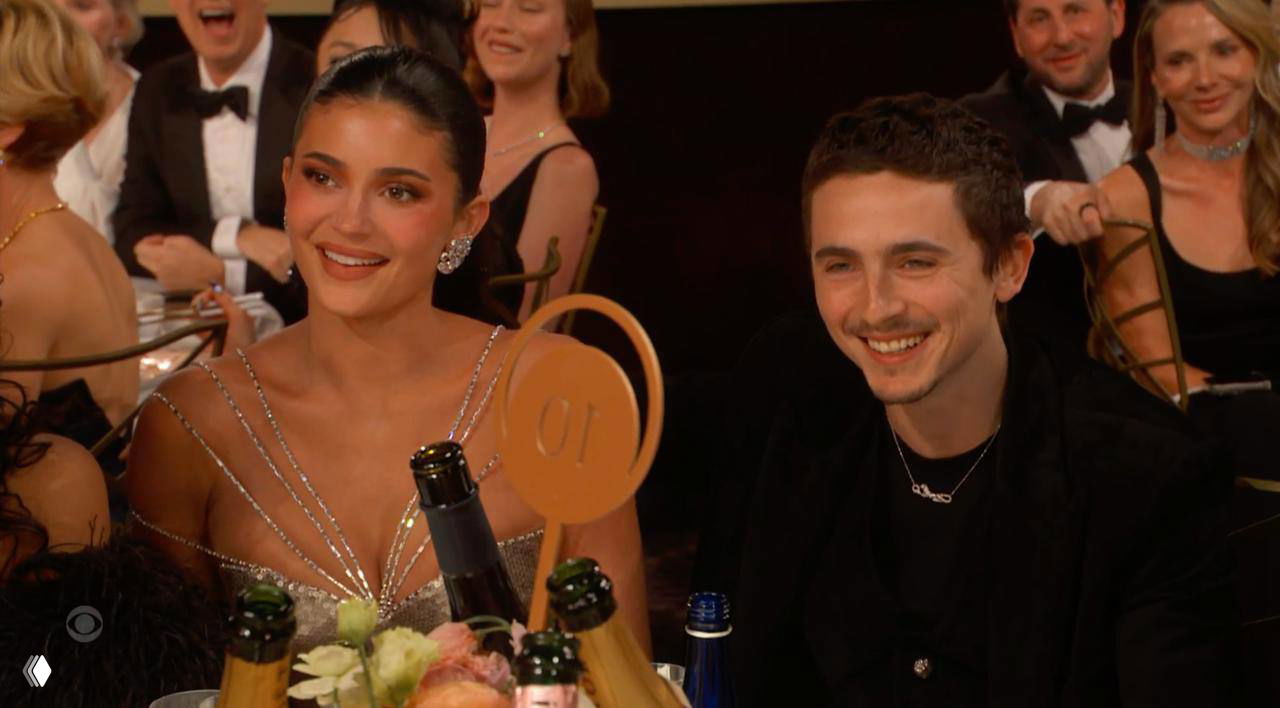 Пара улыбается, сидя за столом на церемонии; на переднем плане цветы и бутылки шампанского, вокруг другие гости в вечерних нарядах.