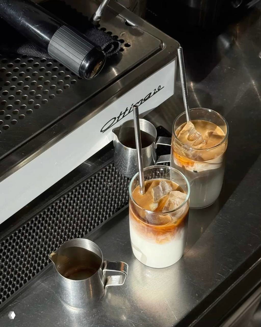 Два айс-латте на металлической барной стойке рядом с кофейным оборудованием и металлическими питчерами, готовые к подаче в кофейне.