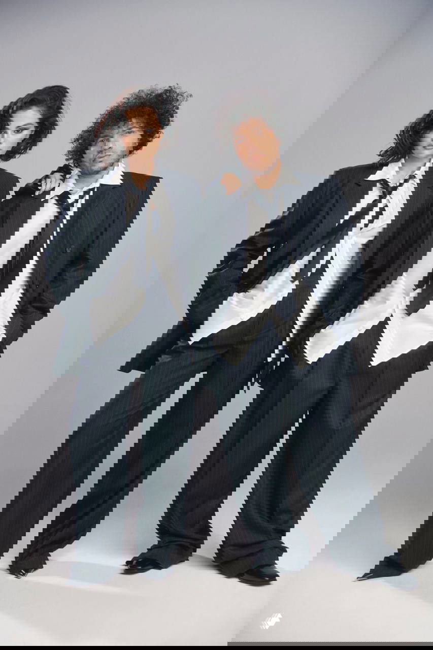 Студийная портретная съёмка двух людей в свободных тёмных костюмах и белых рубашках, нейтральный фон и дружеская поза.