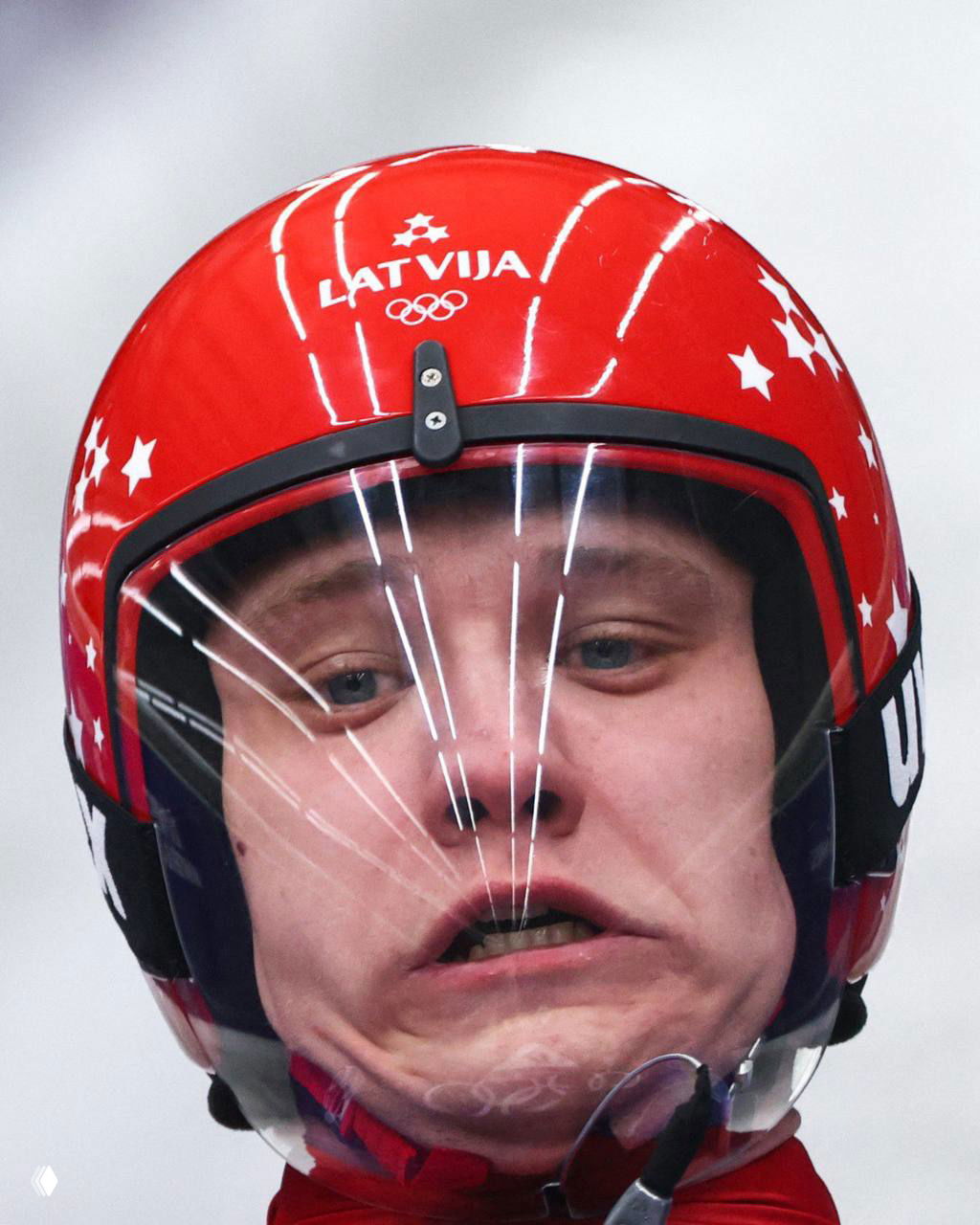 Спортсмен в красном шлеме с надписью LATVIJA, напряжённое лицо и отражения на визоре — необычный и комичный момент на трассе зимних игр.