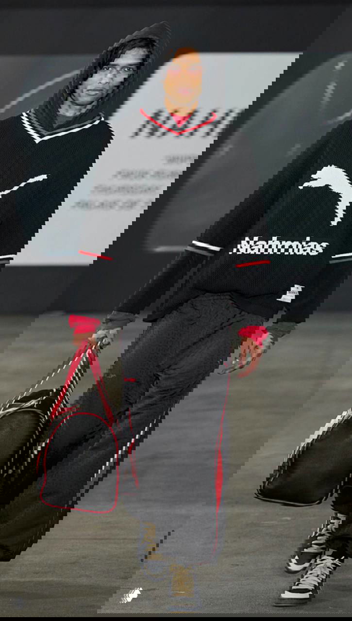 Показ спортивной тематики: модель в тёмном вязаном свитере с логотипом и свободных спортивных брюках, спортивный ретро‑футуризм на подиуме.