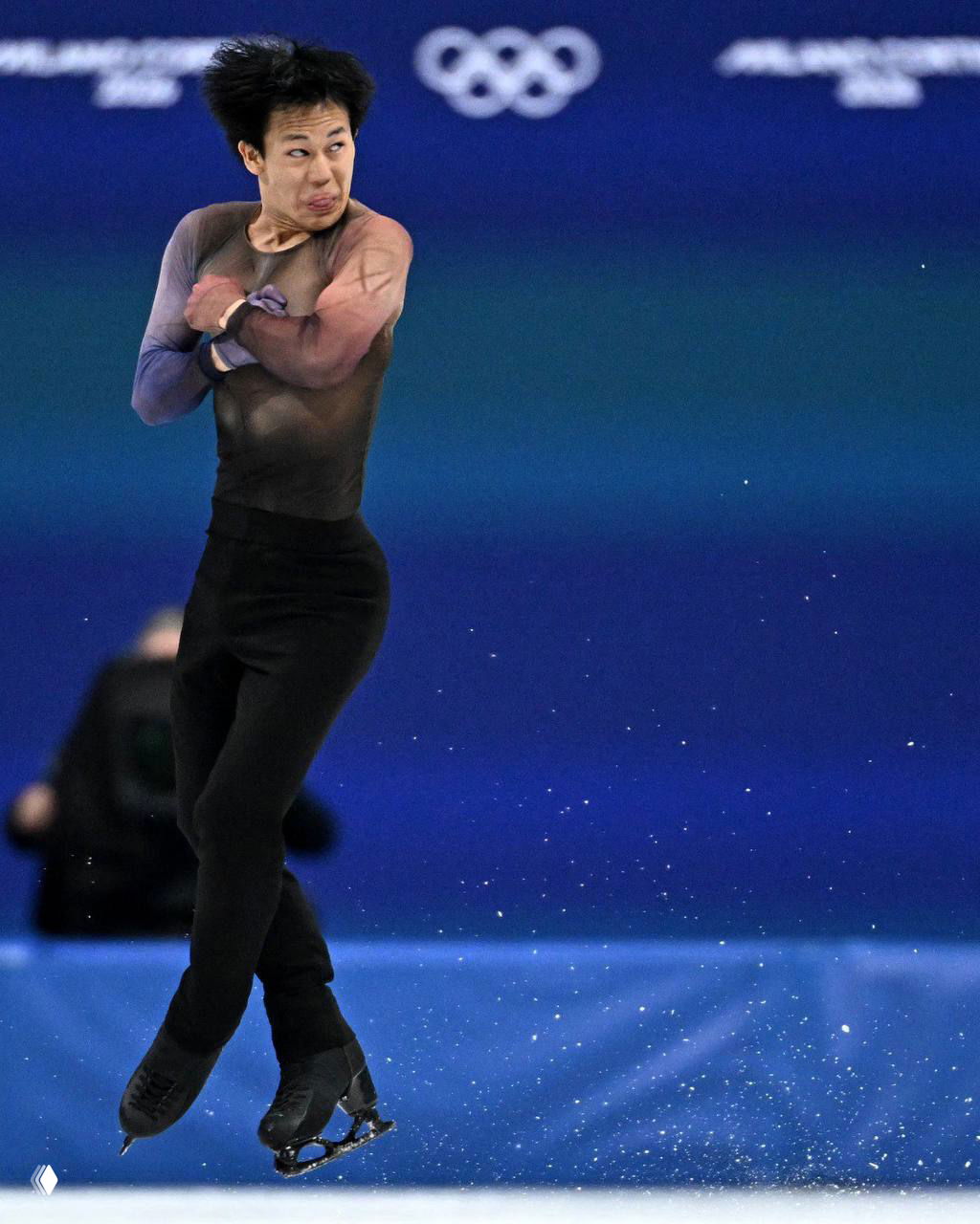 Фигурист на льду с выразительной гримасой и высоким прыжком на фоне олимпийских колец — неловкий, но характерный спортивный кадр.