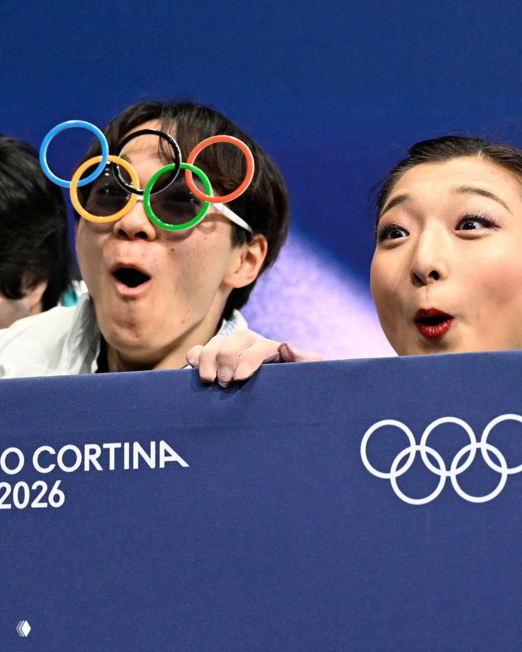 Болельщики на трибуне в очках с олимпийскими кольцами и удивлёнными выражениями лиц — весёлая реакция публики на соревнованиях.