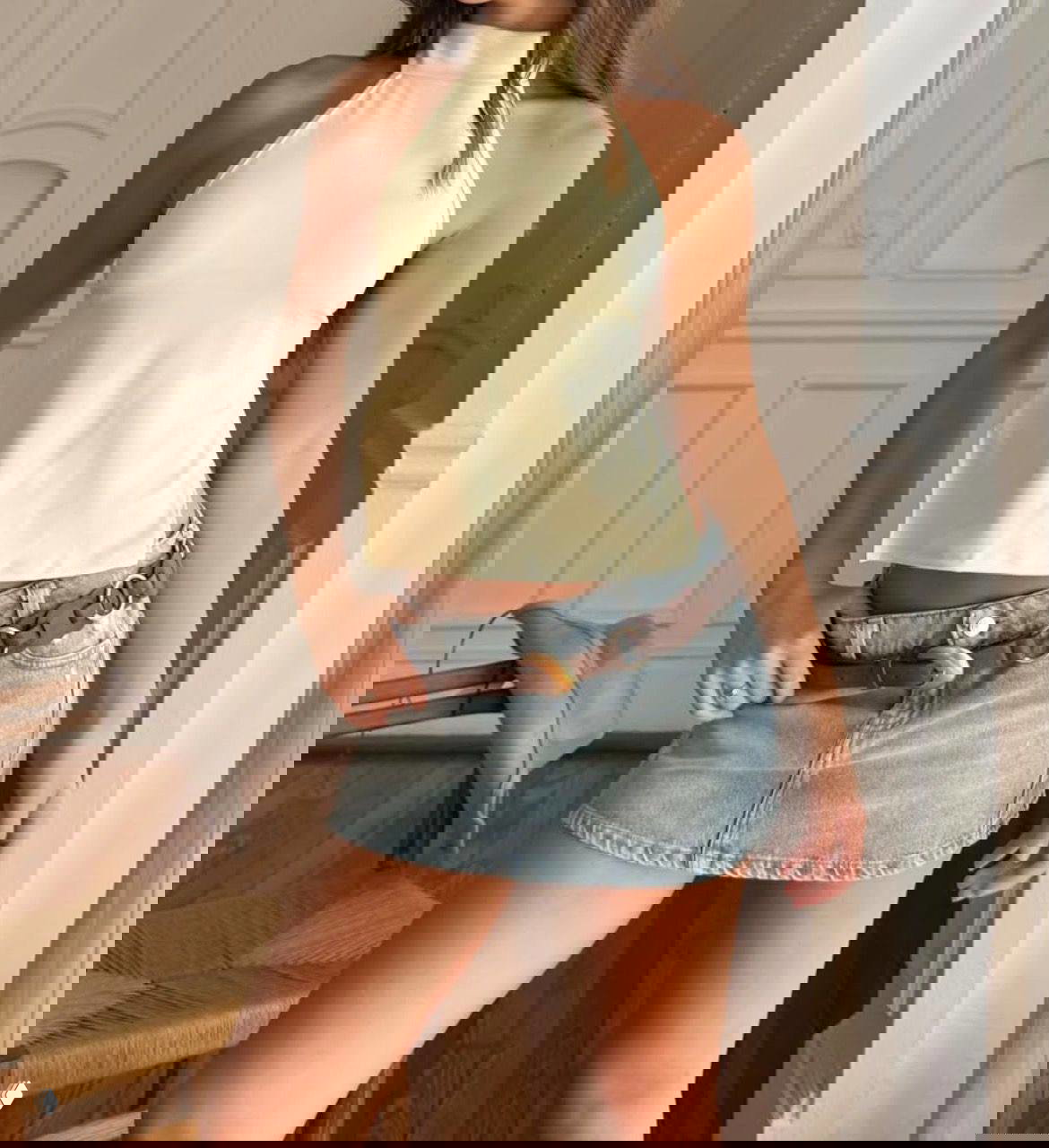 Интерьерное фото: модель в кремовом топе и светлой джинсовой мини-юбке с декоративным ремнём, позирует в комнате с креслом.