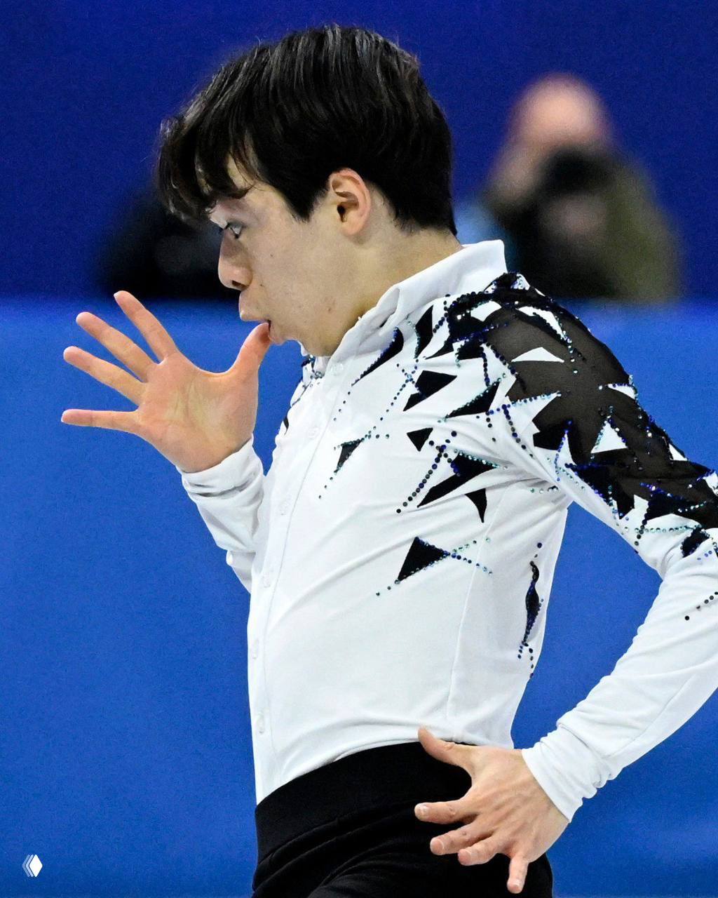 Фигурист в бело‑чёрном костюме делает эффектный жест рукой у рта во время проката, выразительное и немного забавное фото с арены.