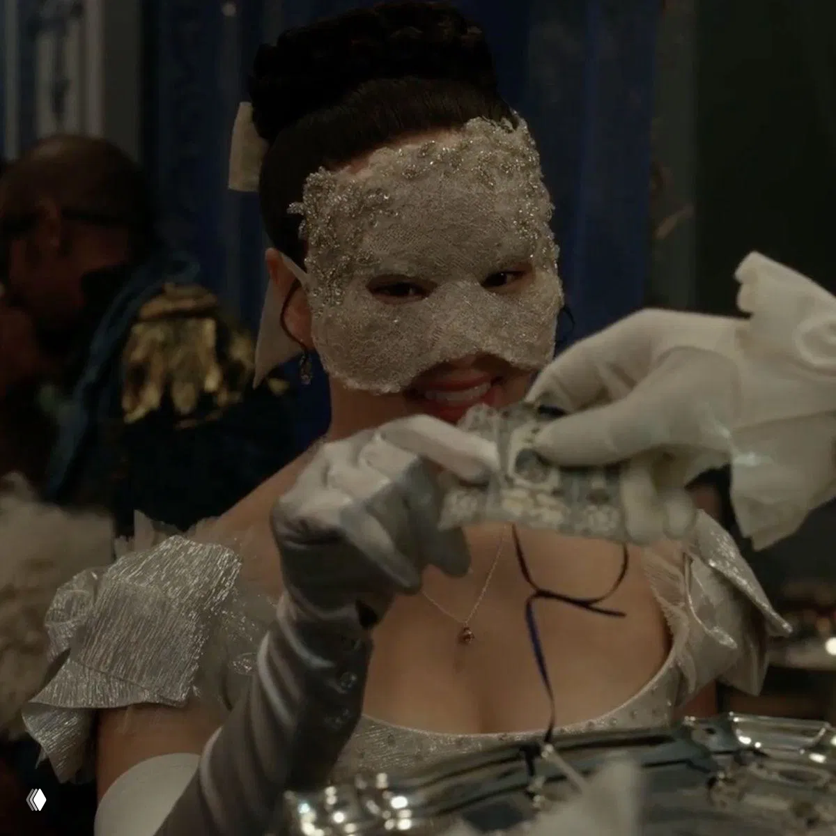 Крупный план героини в декоративной блестящей маске и перчатках, она держит предметы и улыбается в кадре бала.