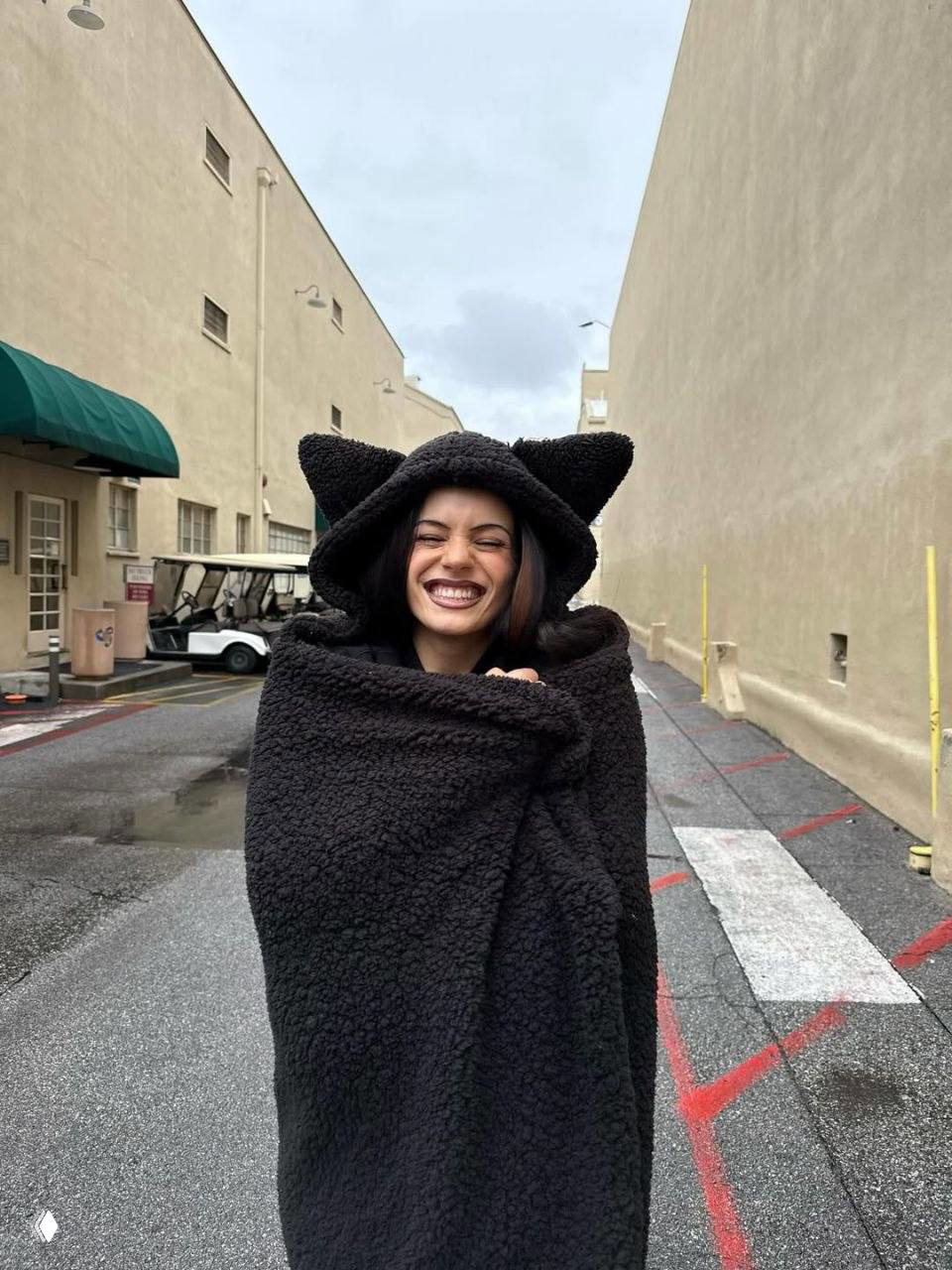 Улыбающаяся женщина, завернутая в тёплый плюшевый плед с ушками, на улице съёмочной базы — повседневный закулисный момент между дублями
