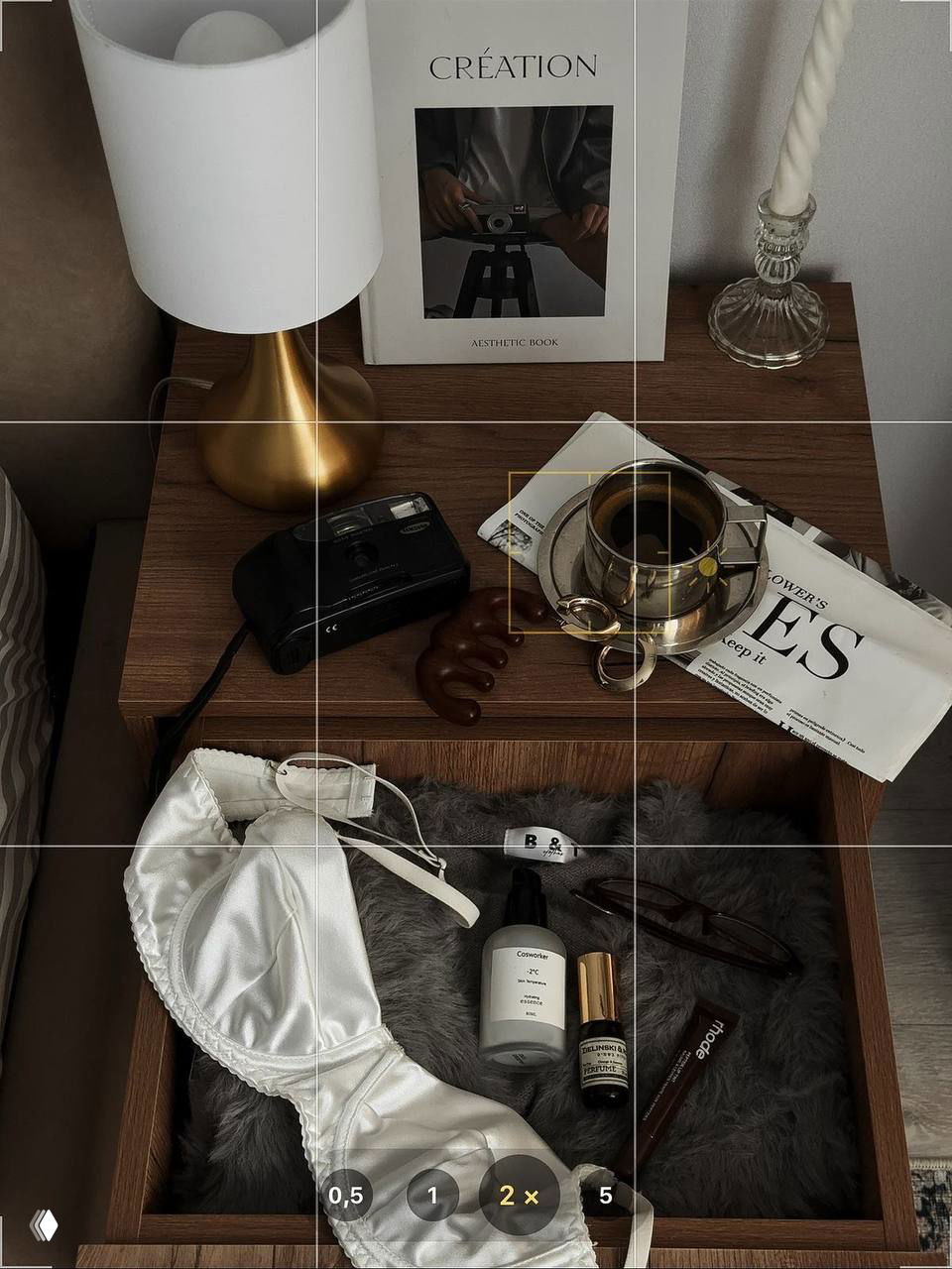 Прикроватная тумбочка с лампой, чашкой кофе и открыенным ящиком с косметикой; интерьерная деталь в нейтральных коричнево-бежевых тонах.