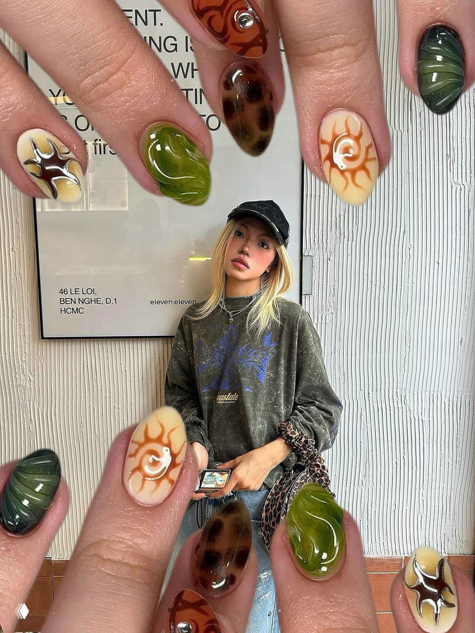 Маникюр с зелёными и коричневыми глянцевыми элементами и рисунком солнца на ногтях в коллаже на фоне девушки в чёрной кепке и свободном свитере.