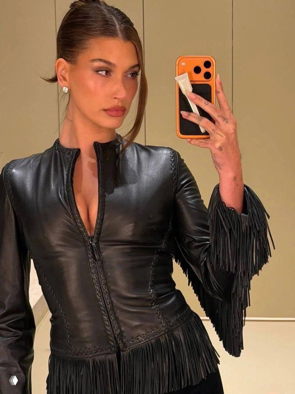 Крупный портретный кадр-селфи Хейли Бибер в зеркале: кожаная верхняя часть с бахромой, аккуратная прическа, заметен маникюр и смартфон в руке.