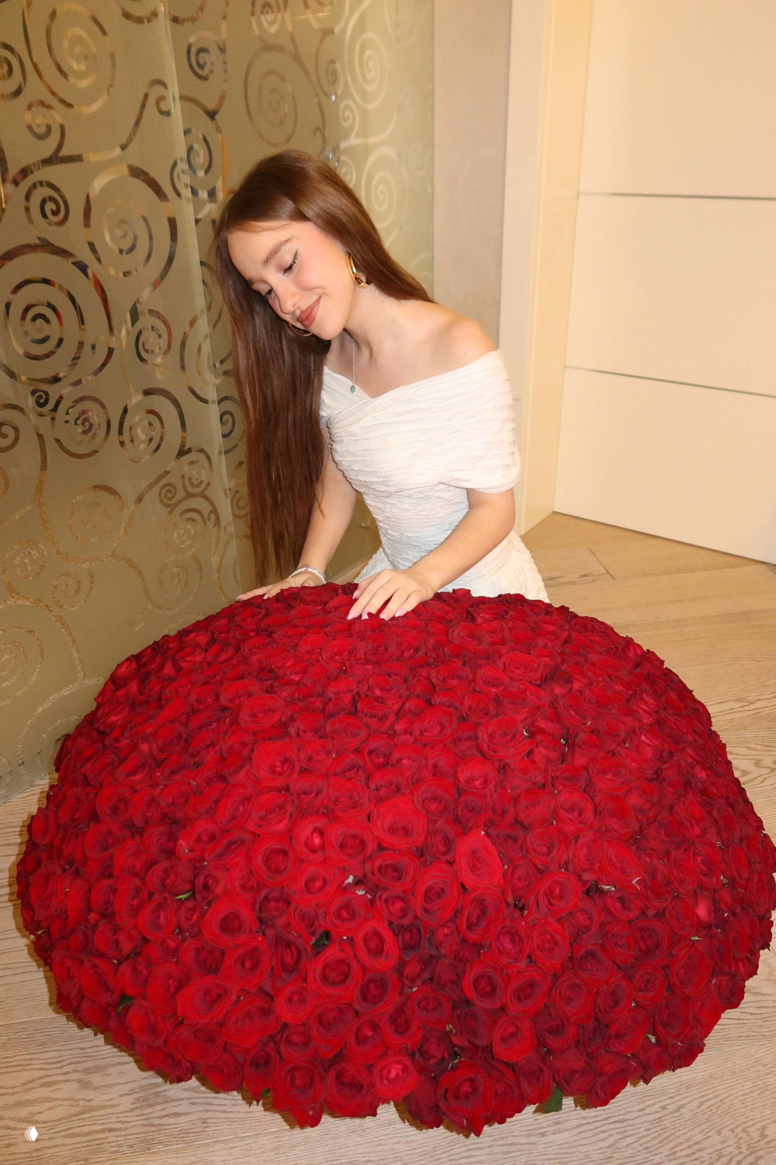 Марьяна Локель в белом платье прислонилась к большой куполообразной композиции из 1001 красной розы на полу — общий план.