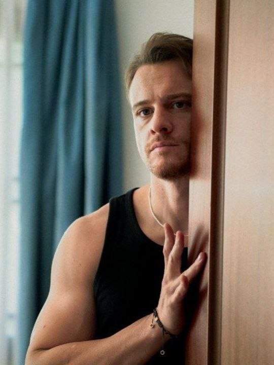 Портрет Керем Бюрсина: мужчина с лёгкой щетиной и серьёзным взглядом выглядывает из дверного проёма, кинематографичный кадр.