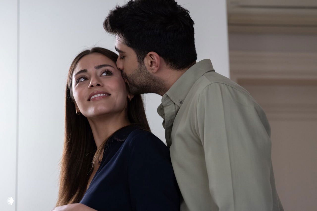 Кадр с тёплым жестом: мужчина целует женщину в щёку, оба выглядят задумчиво — момент близости между персонажами.