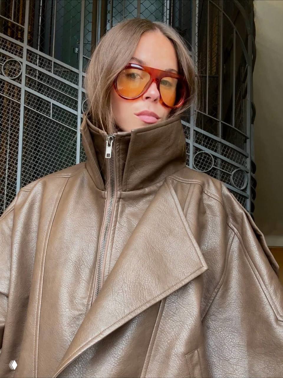Портрет девушки в больших янтарных очках oversize и коричневой кожаной куртке: винтажный силуэт рамки, подчёркнутый высокий ворот, интерьерная арка на заднем плане.