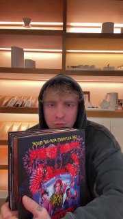 Человек в худи крупным планом держит книгу/пластинку перед камерой на фоне полок с товарами, нейтральное выражение.