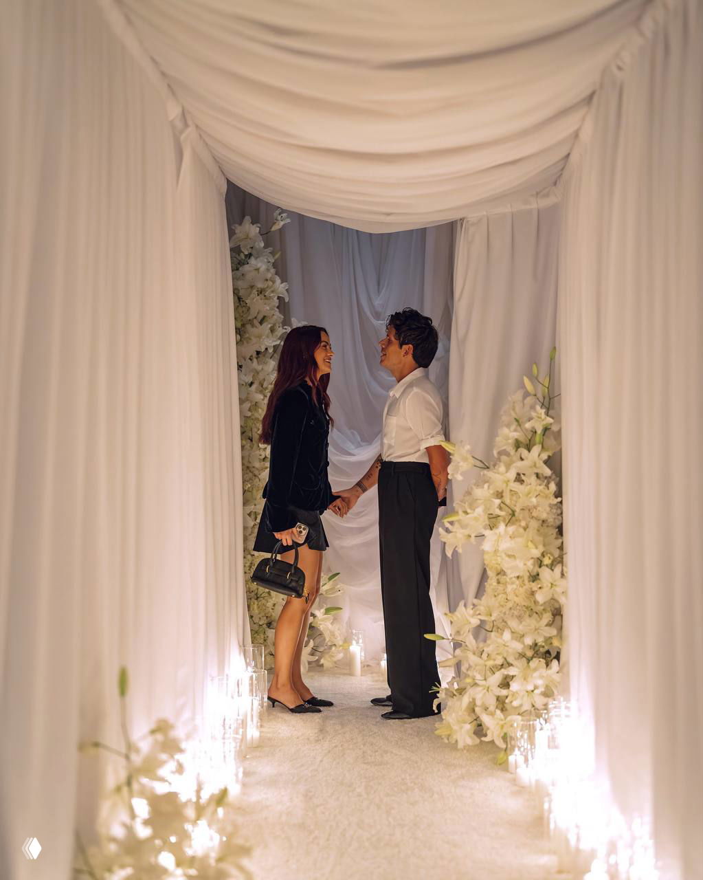 Камила Мендes и Руди Манкузо стоят лицом друг к другу, держат руки в украшенном цветами белом проходе с подсветкой