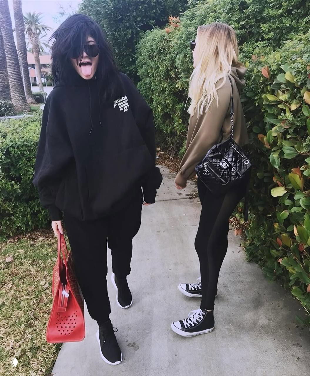 Две девушки прогуливаются по тротуару в повседневной одежде и худи, одна показывает язык — уличный кадр с непринуждённым настроением 2016–2018.