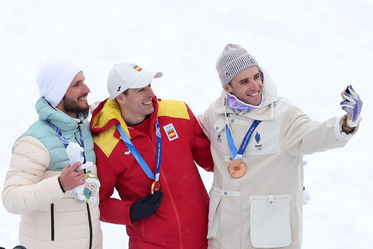 Три спортсмена на церемонии награждения в зимней форме на заснеженной арене: один в красной куртке держит медаль, все улыбаются и делают селфи на снегу.