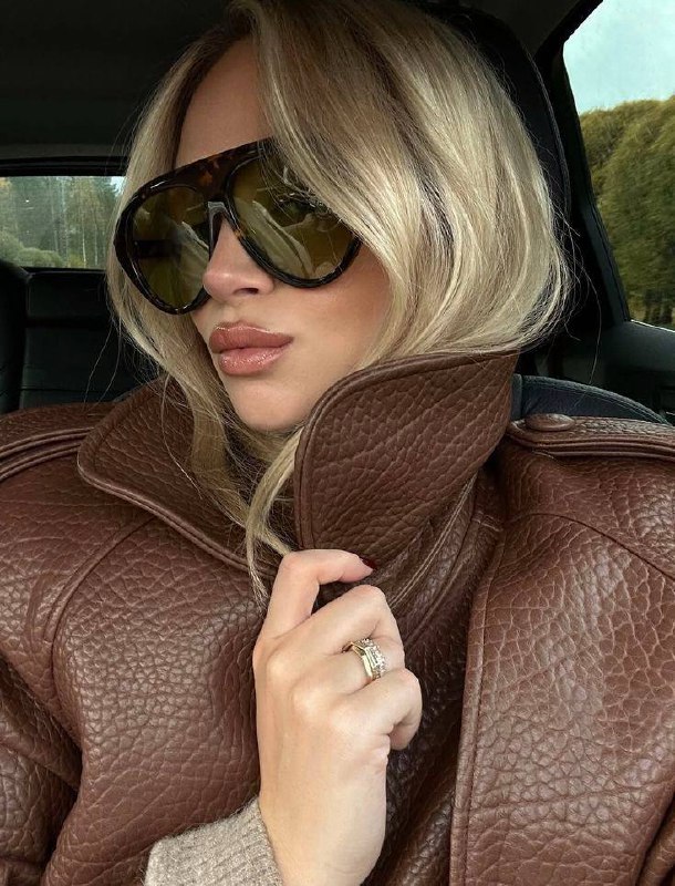 Крупный план женщины в объёмных солнцезащитных очках oversize в автомобиле: тёмно-коричневая оправа, глянцевая текстура кожи пальто и отражения в линзах.