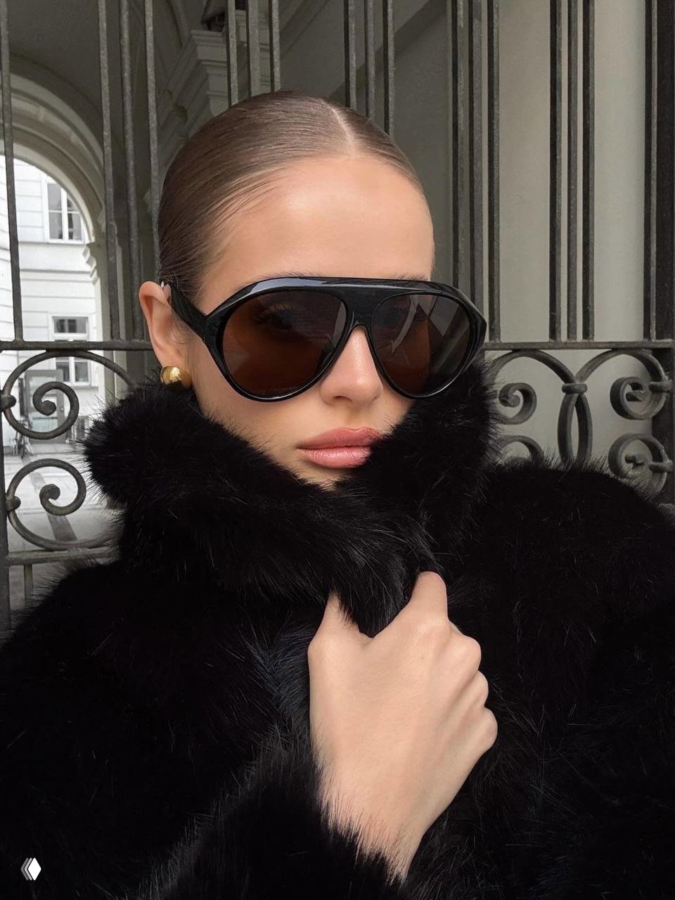 Модель в чёрных больших очках oversize и чёрной шубе с высоким воротником: гламурный зимний образ, строгий макияж и минималистичная архитектура на фоне.