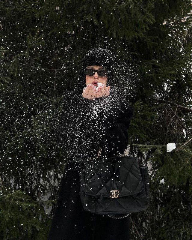 Девушка в тёмном пальто и очках разбрасывает снег руками перед хвойным фоном — динамичный зимний кадр с чёрной сумкой-«квинтэссенцией» образа.